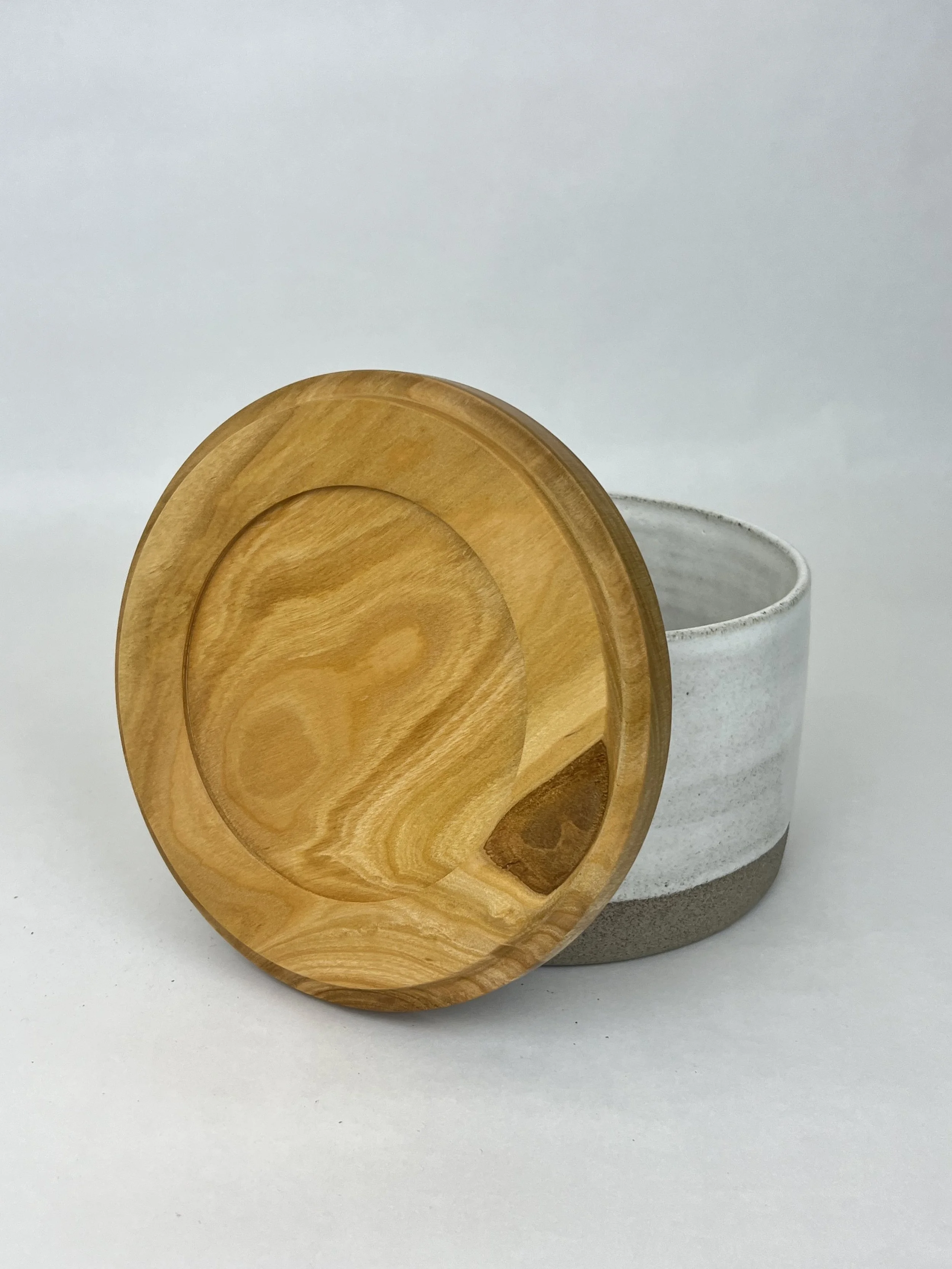Eine weiße Tasse mit einem grauen und weißen Muster, daneben ein runder Holzdeckel mit einer natürlichen Holzmaserung.