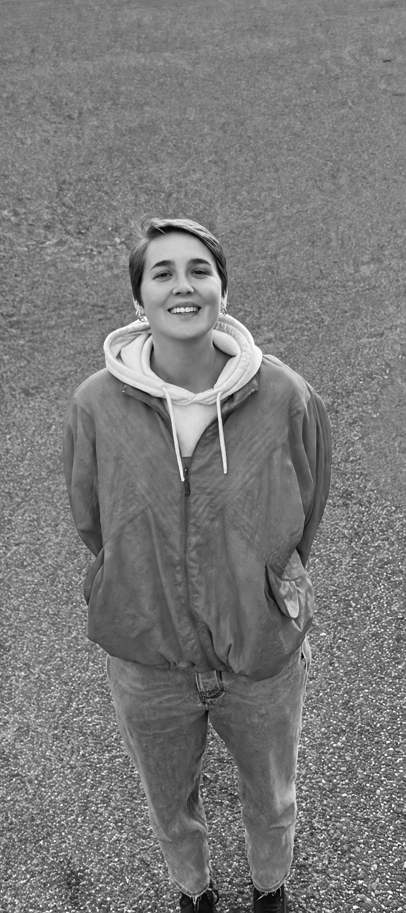 Eine junge Frau mit kurzen Haaren, lächelnd, trägt eine Joggingjacke und eine Kapuzen-Sweatshirt, steht auf einer Asphaltstraße.