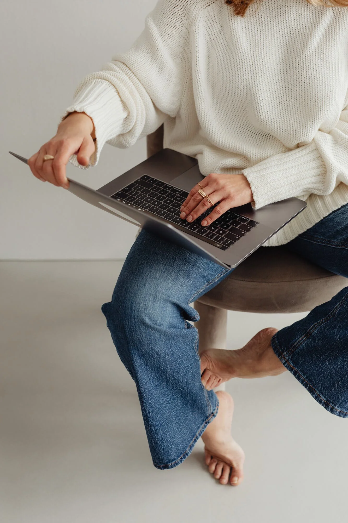 woman in jeans and white sweater sitting on a stool and typing on her laptop