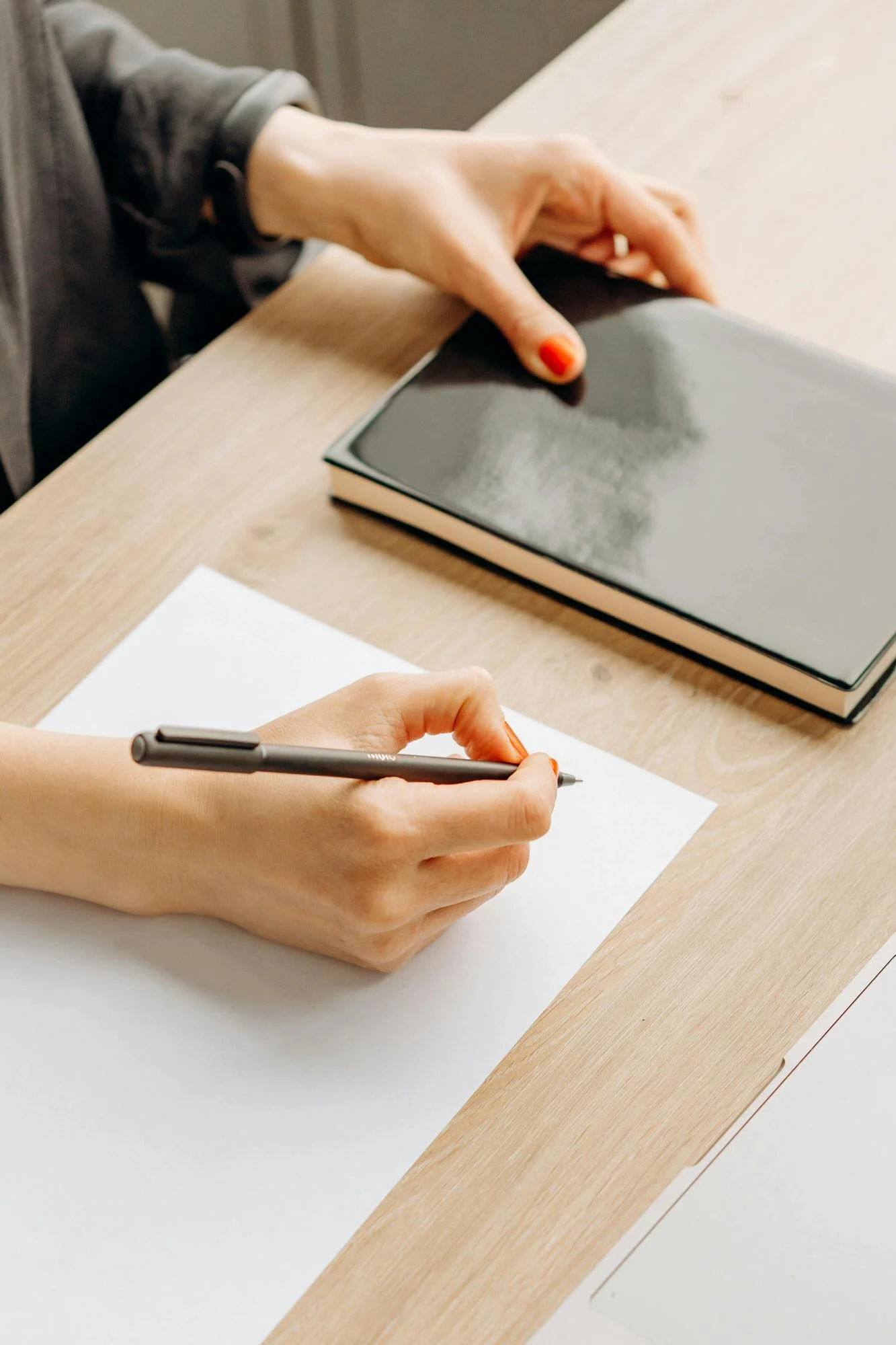 woman holding a pen and writing on a piece of white paper on tan table top with a black hardcover book next to her