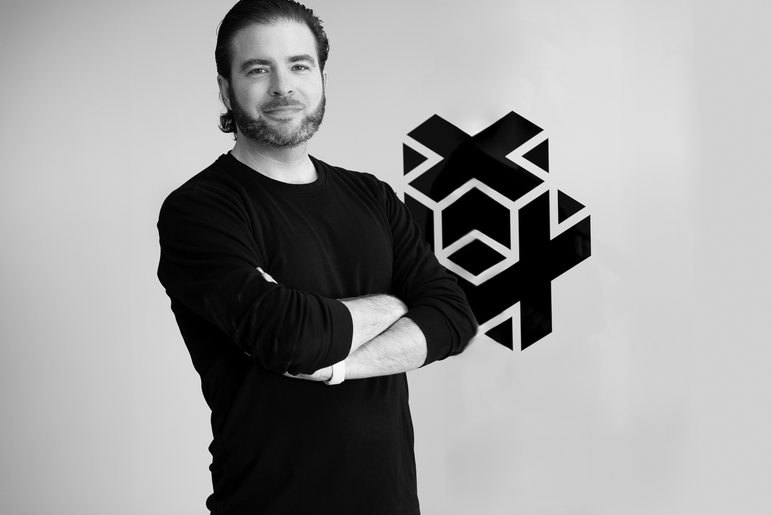 A man with dark hair and a beard, wearing a black long-sleeve shirt, standing with arms crossed in front of a geometric logo on a wall.