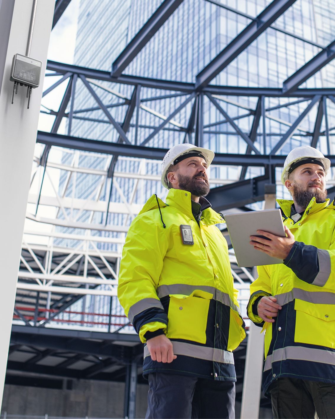 Two construction workers wearing yellow jackets and white safety helmets standing inside a modern building with glass and steel architecture, looking up and discussing construction plans.