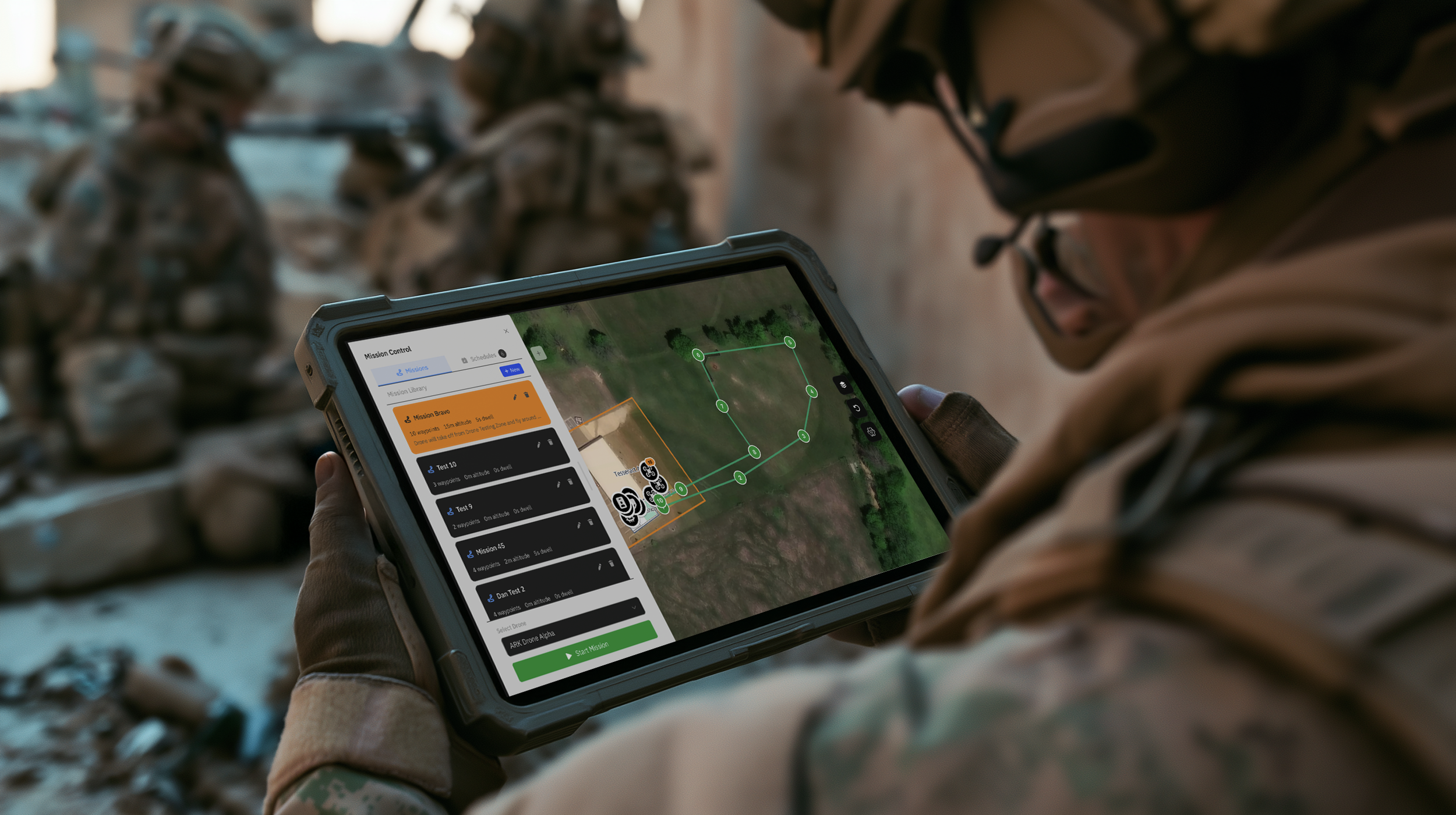 A soldier in a camouflage uniform holding a rugged tablet displaying a drone mission-planning Site Manager interface, with a group of soldiers blurred in the background.