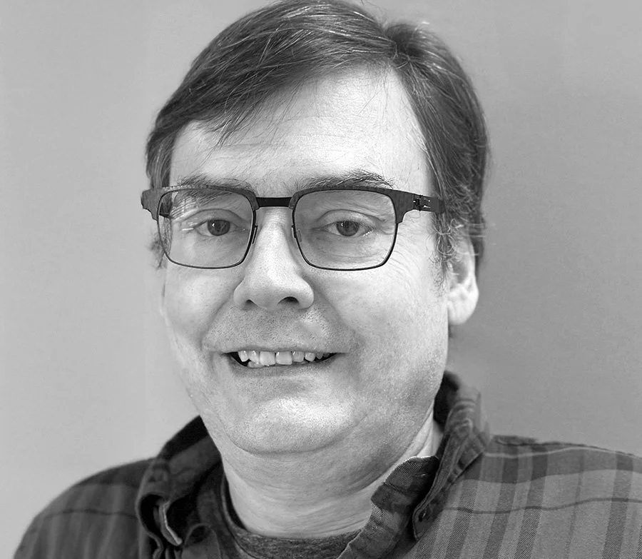 Black and white portrait of a smiling man with glasses, wearing a checked shirt, in front of a plain background.