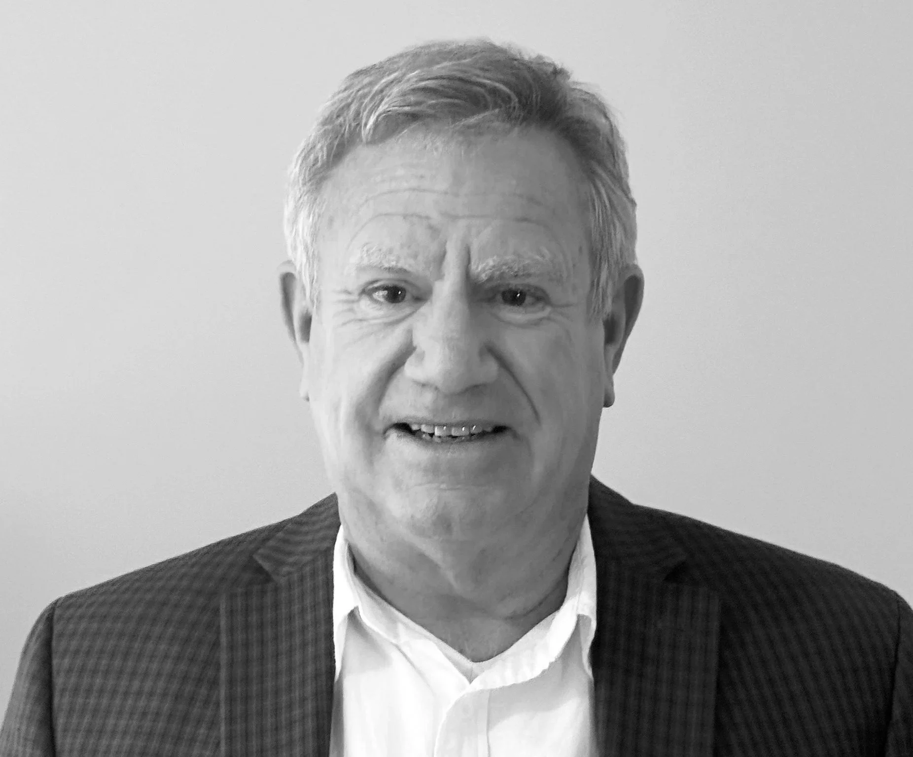 A black-and-white photograph of a man with short, combed-back hair, wearing a plaid suit jacket and a white shirt. He is smiling slightly and looking directly at the camera, with a plain wall background.