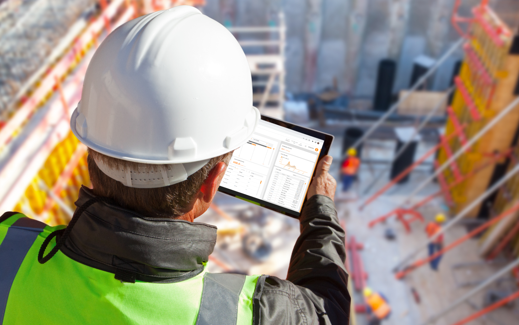 Construction worker wearing a white safety helmet and high-visibility vest holding a tablet with PRISM Dashboard data charts on a construction site.