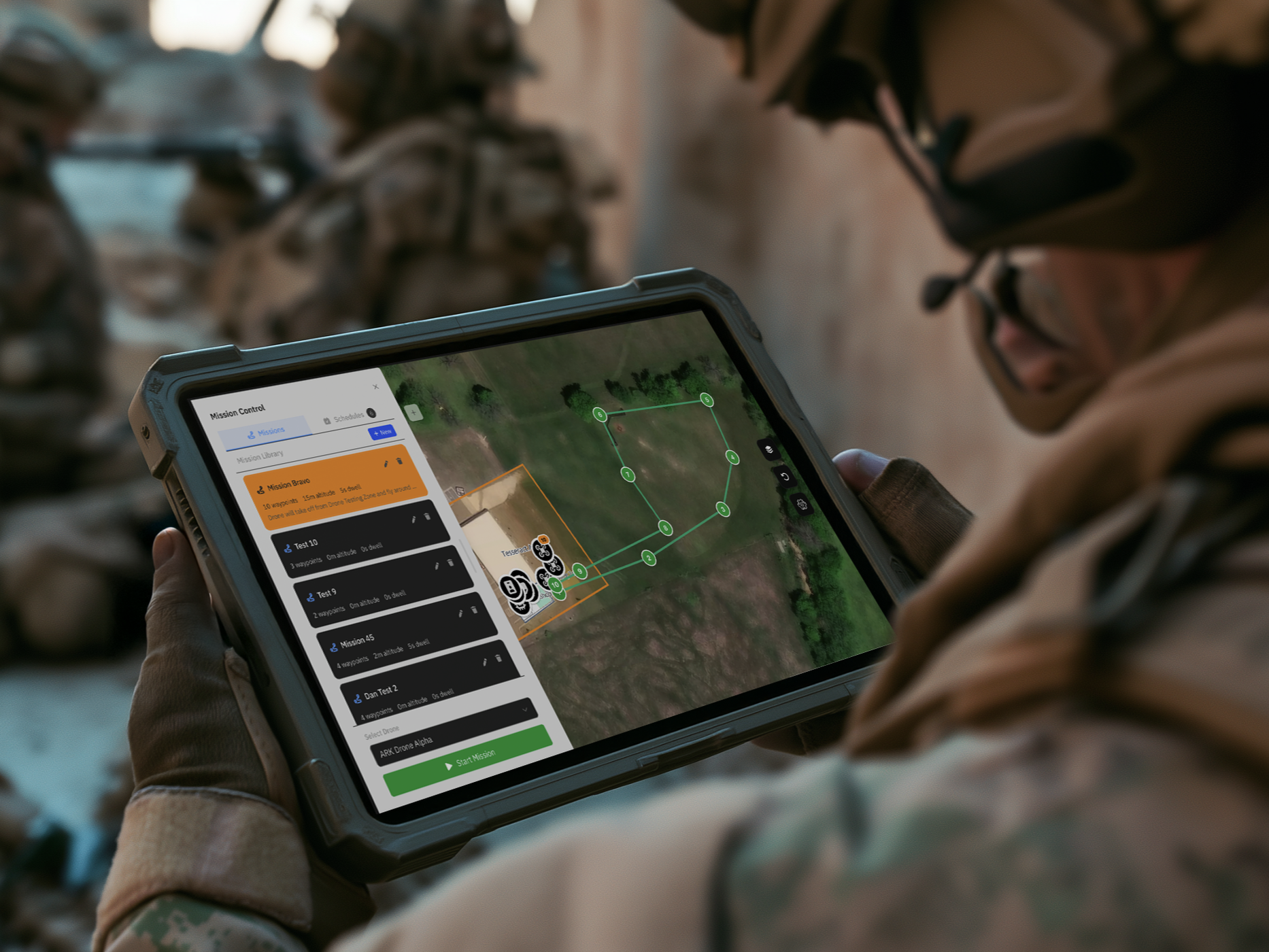 A person in military camouflage uniform holding a rugged tablet with a drone mission planning app displayed, showing a map, mission details, and controls, likely outdoors with trees and military gear in the background.