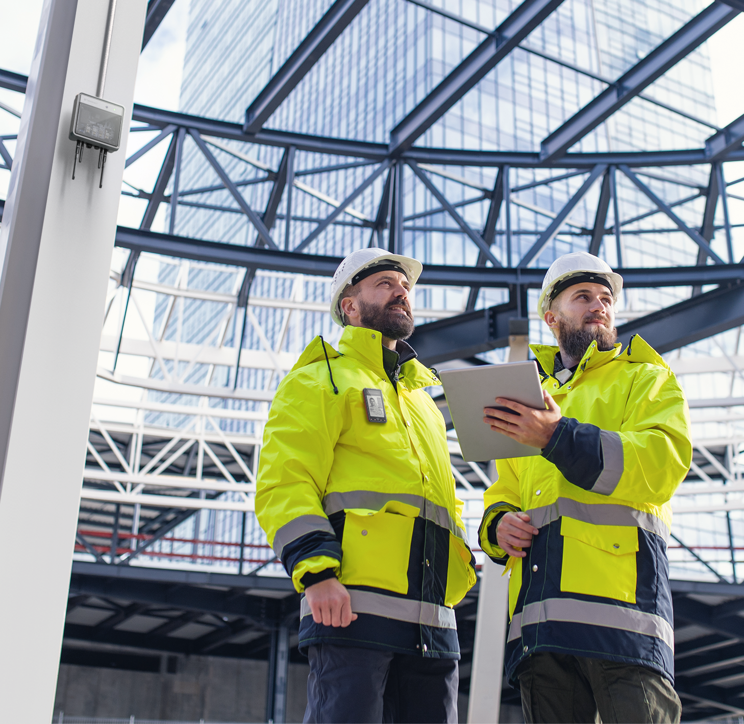 Two construction workers in yellow safety jackets and white helmets inspecting equipment inside a modern building with glass walls and a steel framework.