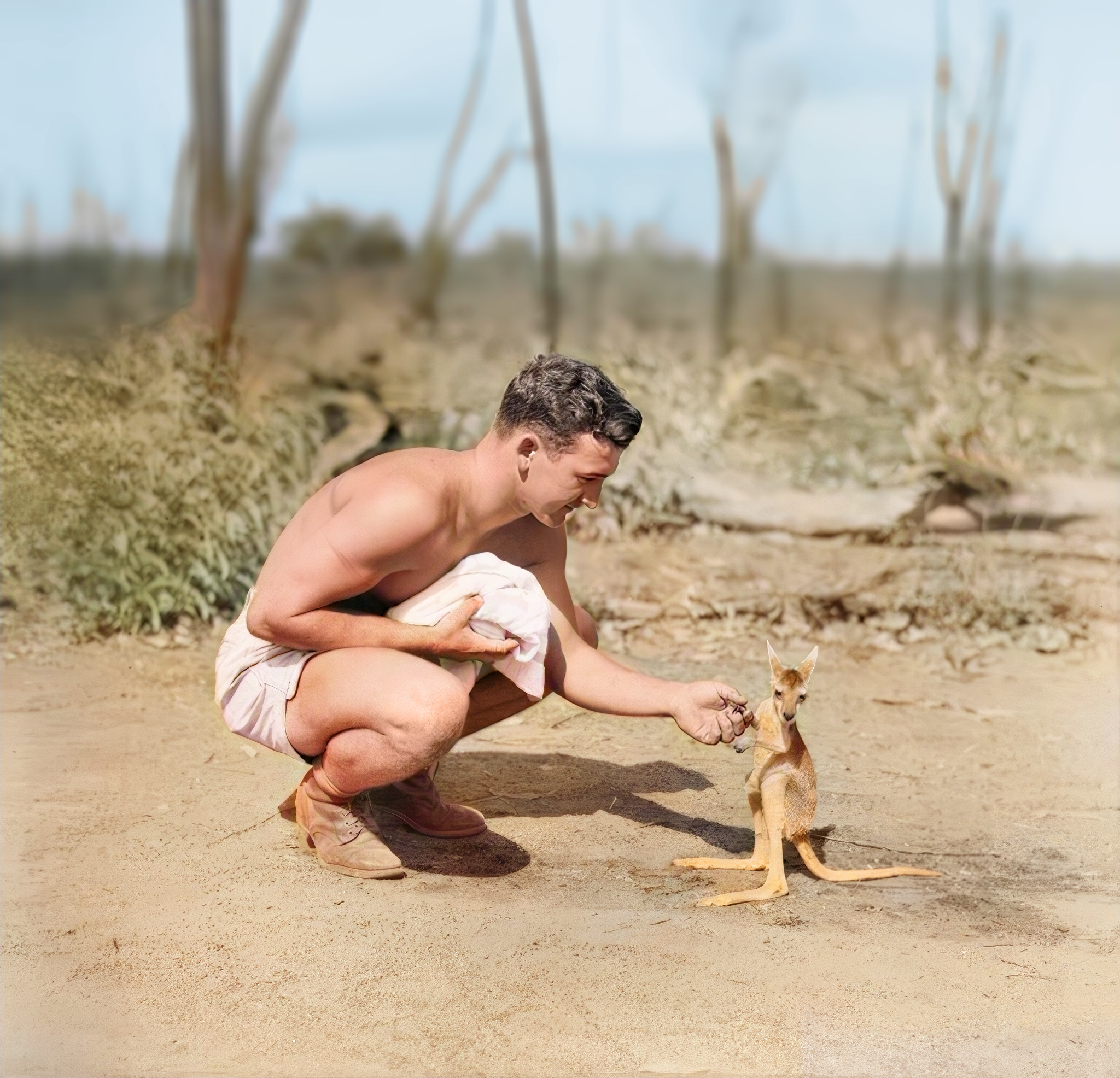 A young man squatting on the ground outdoors, holding a small animal, possibly a kitten, in his hands. The background features dry, barren land and sparse trees.