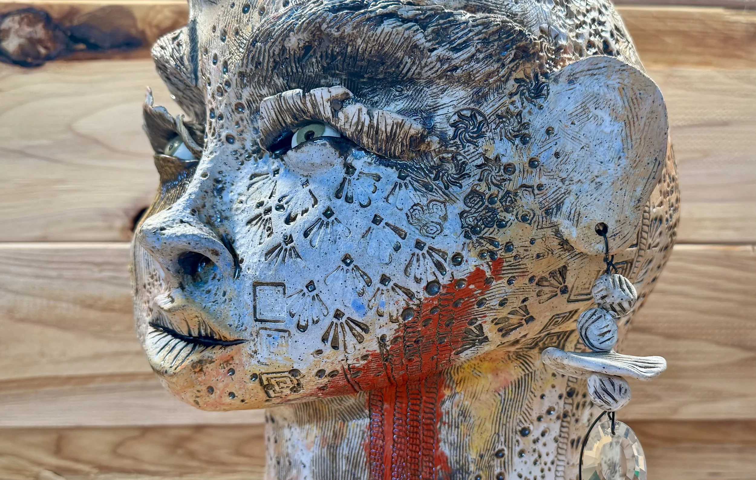 A sculpture of a human face with intricate carvings and patterns, including a prominent nose, partially open eyes, and a red stripe running down the mouth area, set against a wooden background.