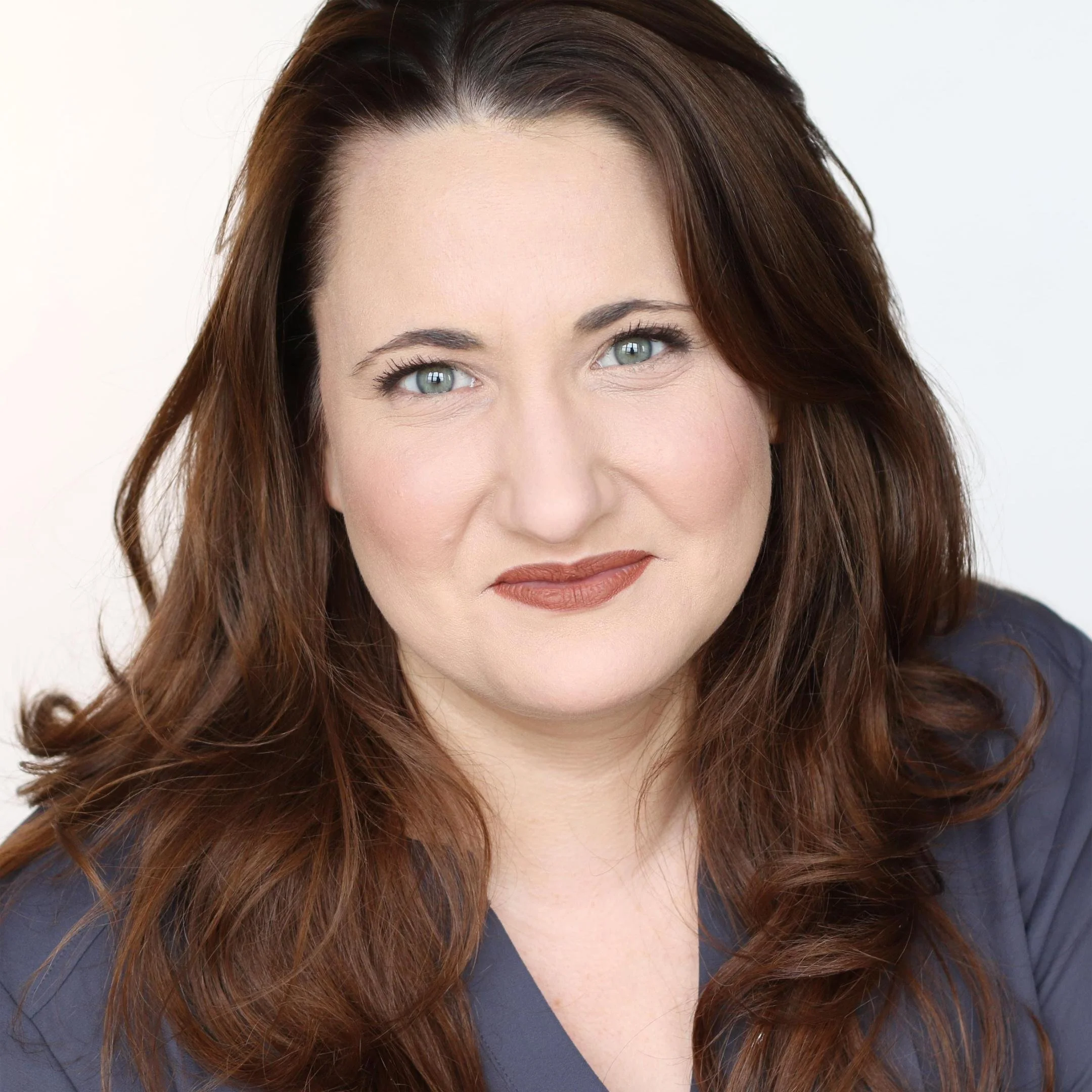 Close-up of a woman with brown hair, blue eyes, wearing light makeup, and a dark top, against a plain white background.