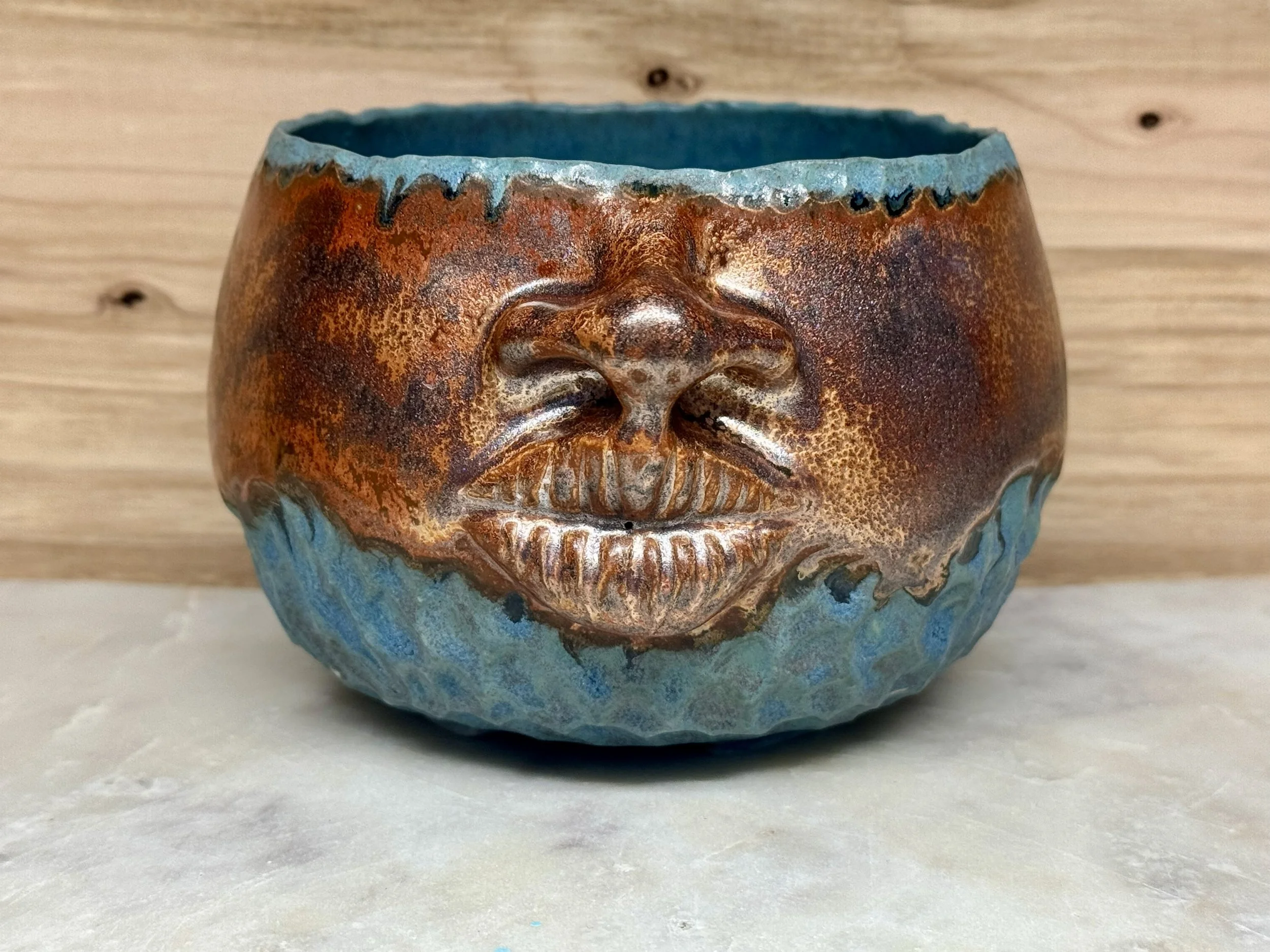 A ceramic cup with a face design, featuring closed eyes, a nose, and a smiling mouth, with a textured surface and a blue and copper glaze finish.