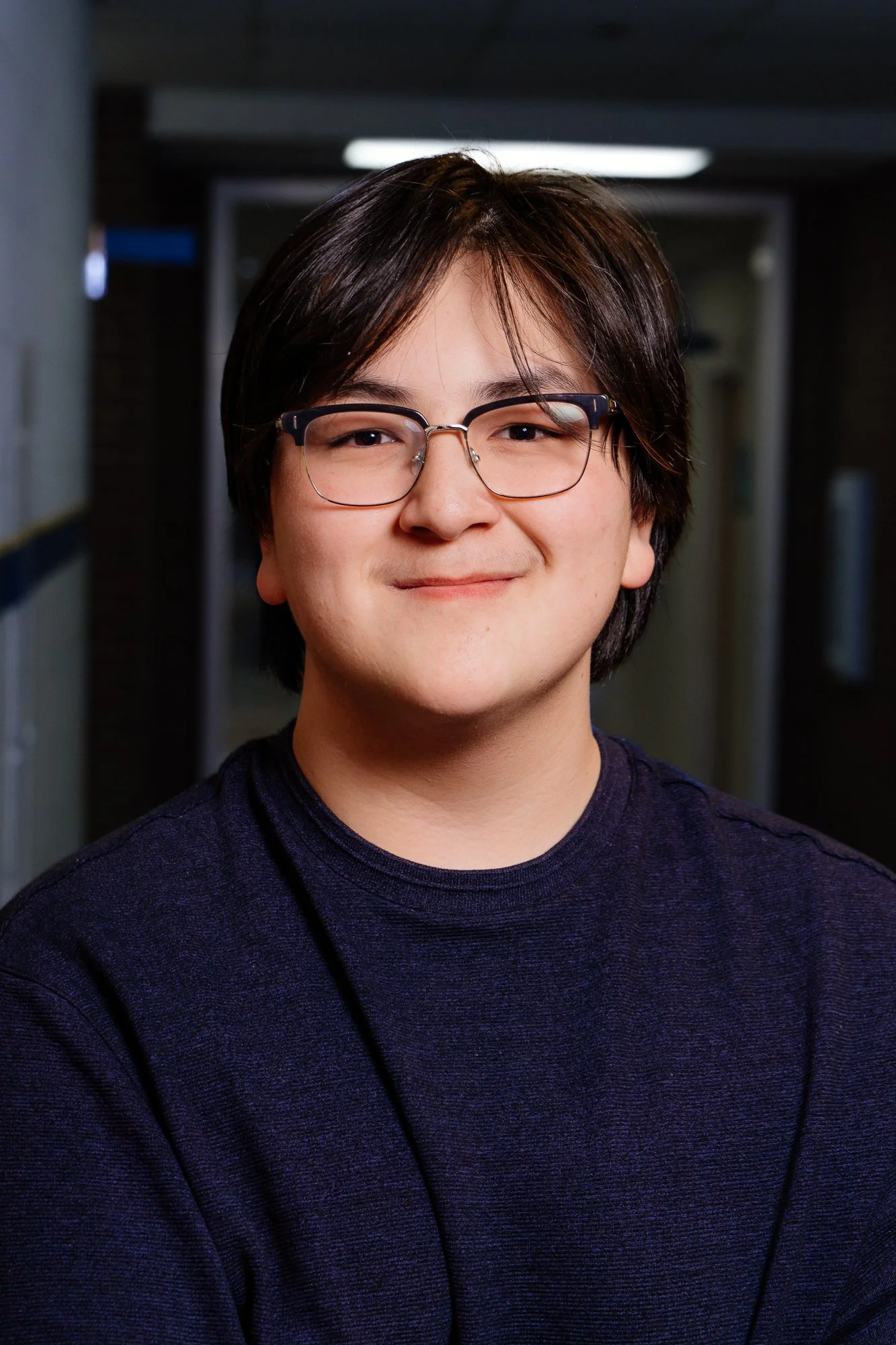 Young man with dark hair, glasses, and a slight smile, standing in a hallway.
