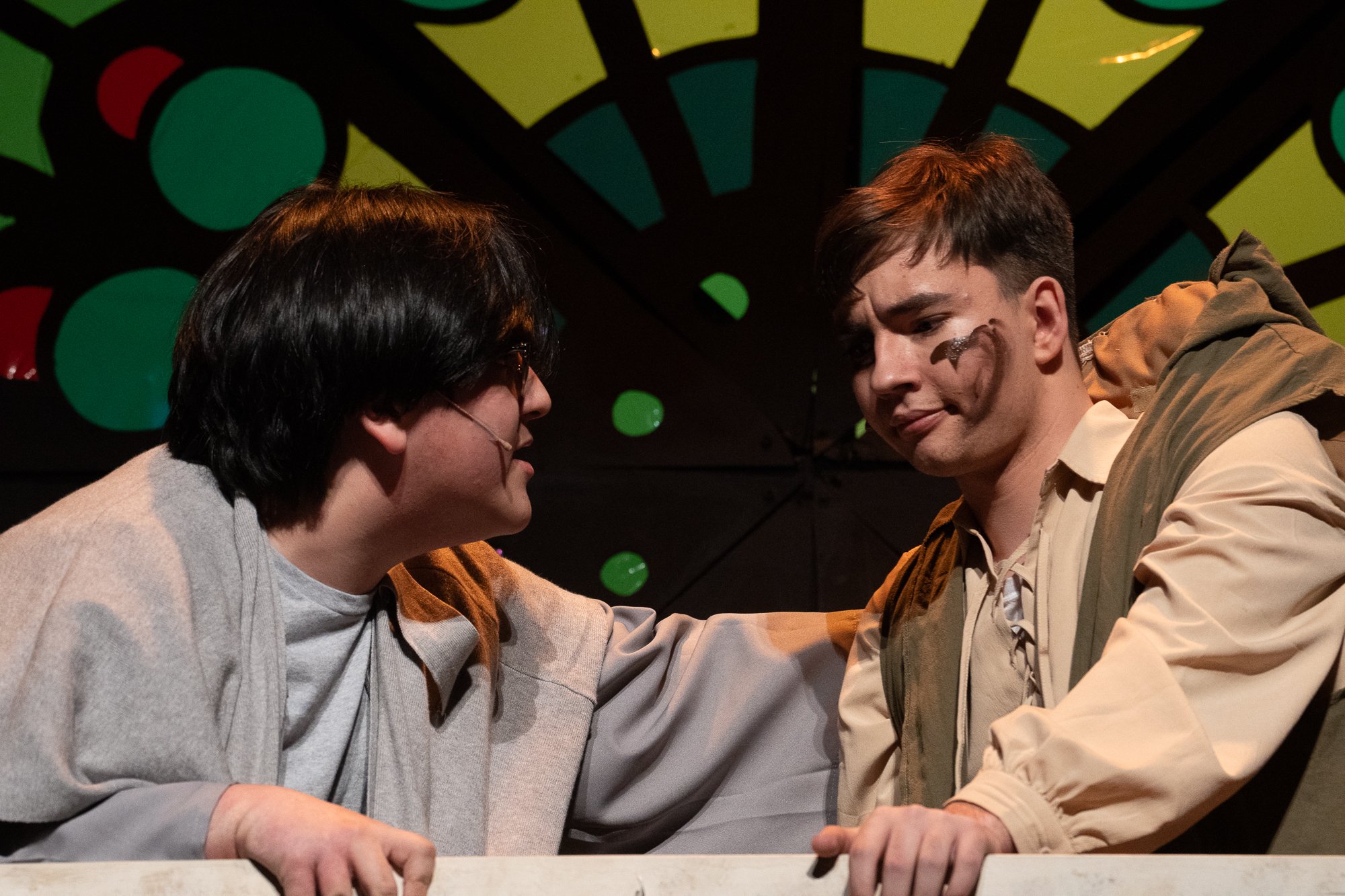 Two young men are depicted in close-up, leaning towards each other. One has dark hair, glasses, and is smiling, while the other has light brown hair, a face painted with a black mark, and appears somber. They are against a colorful, circular, geometric background.
