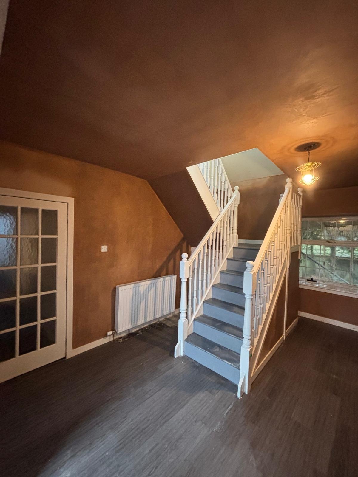 Interior of a house with a staircase ledge and white handrails, a brown wall, a closed glass-paneled door on the left, a white radiator below a thermostat, a window with a grid pattern on the right, and a ceiling light fixture illuminating the space.