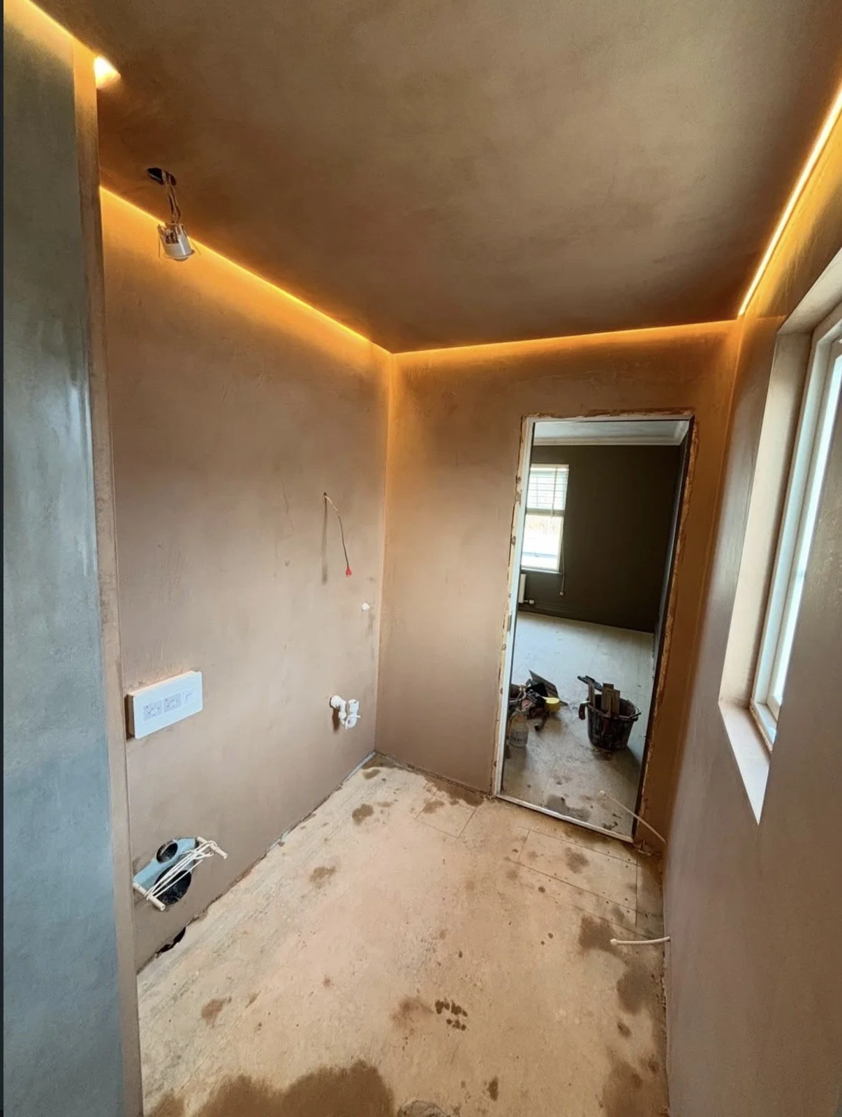 A room under construction with unfinished walls, a window, and exposed wiring. The floor is dirty with construction debris, and there are tools and materials in another room visible through a doorway.