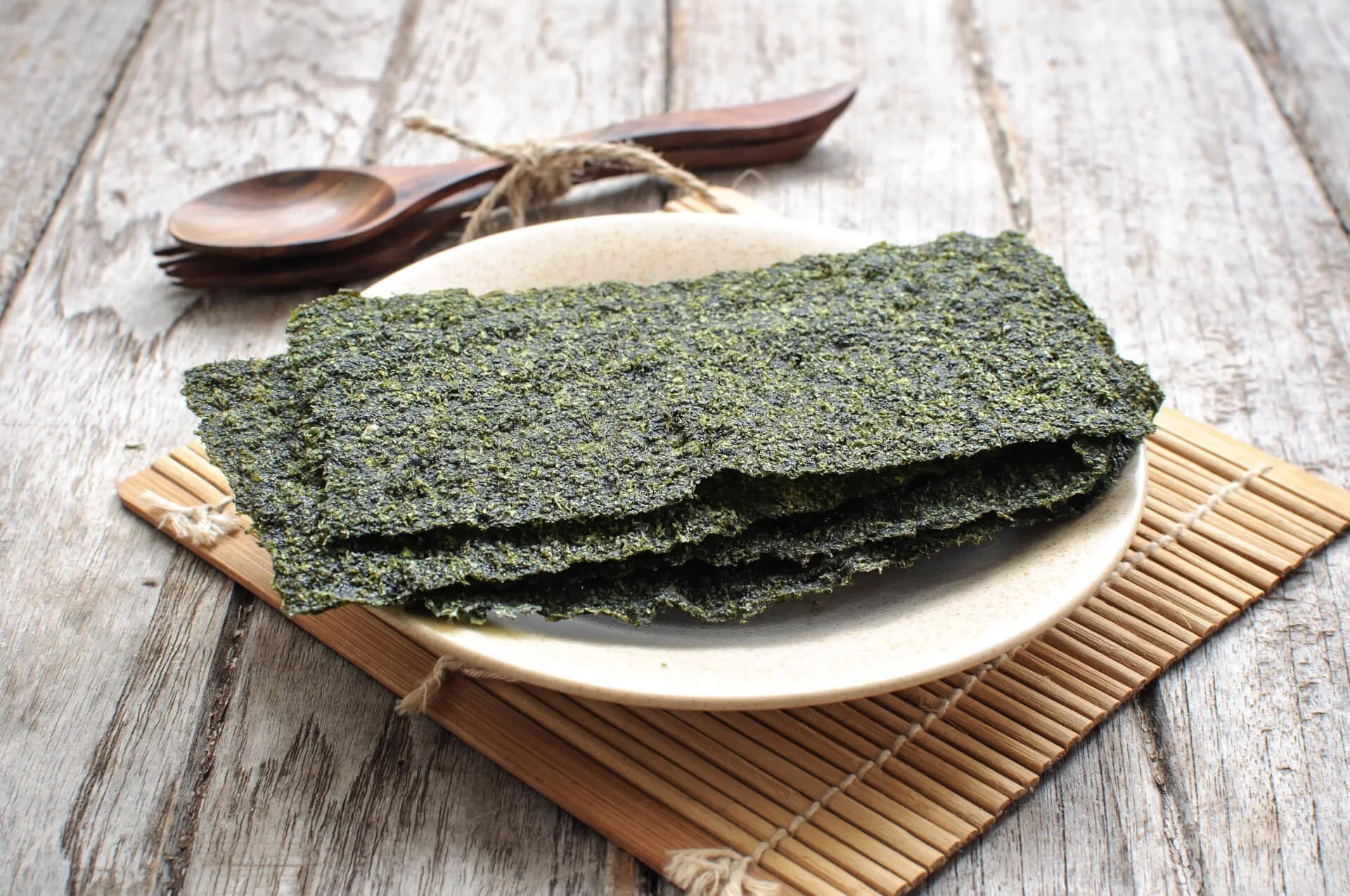 Stack of dried seaweed sheets on a white plate, placed on a bamboo mat with wooden utensils in the background.