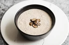 A black bowl of creamy soup garnished with three small pieces of mushrooms, placed on a white background.