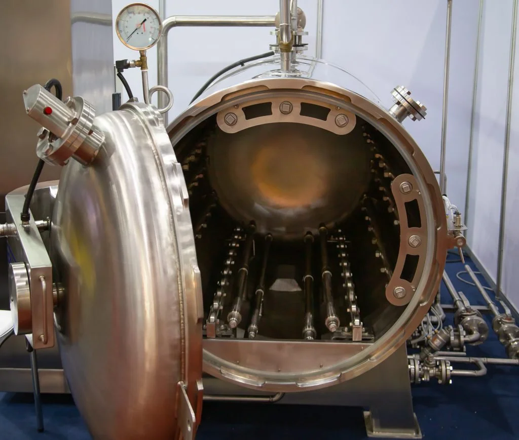 A large stainless steel vacuum chamber with a circular opening, surrounded by bolts and brackets, with attached pipes and gauges for pressure and temperature monitoring, used in scientific or industrial applications.