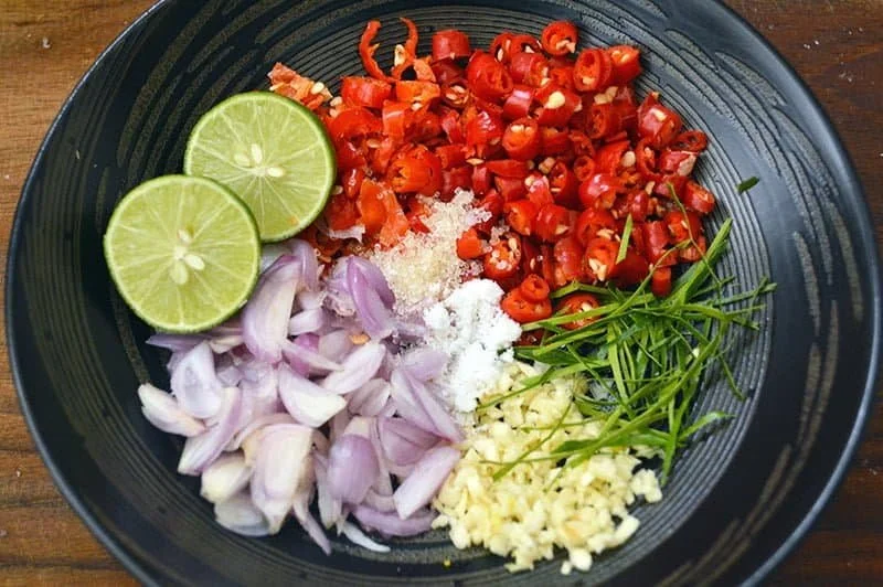 A black bowl containing chopped red chili peppers, two slices of lime, minced garlic, chopped shallots, chopped green herbs, and grated cheese.