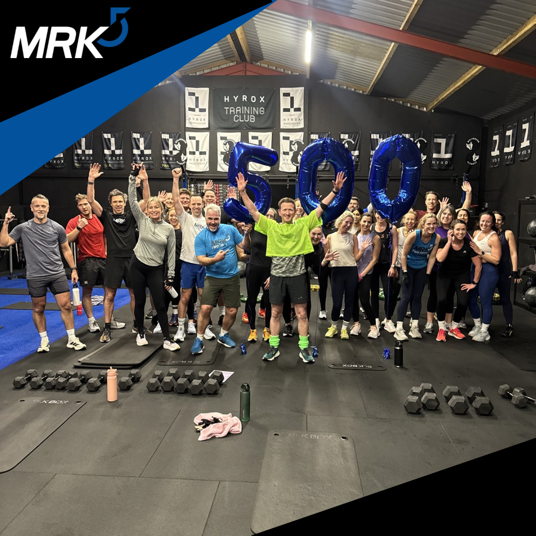 Group of people celebrating in a gym, holding large blue balloons forming '500', with a black banner in the background that reads 'HYROX TRAINING CLUB', and gym equipment in the foreground.