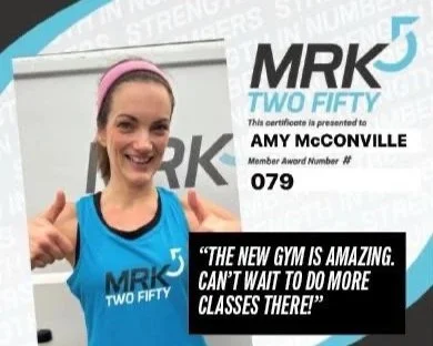 A woman smiling and giving a thumbs up, wearing a blue tank top with 'MRK TWO FIFTY' logo, standing next to a certificate that reads 'MRK TWO FIFTY,' awarded to Amy McConville with member award number 079, and a quote: 'The new gym is amazing. Can't wait to do more classes there!'