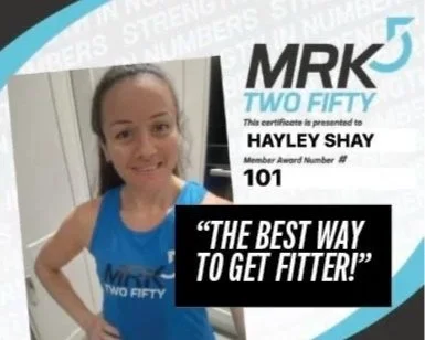 A woman in a blue shirt with a logo, holding a certificate that says 'MRK 50 Two Fifty' and 'Hayley Shay,' with a quote 'The best way to get fitter!'