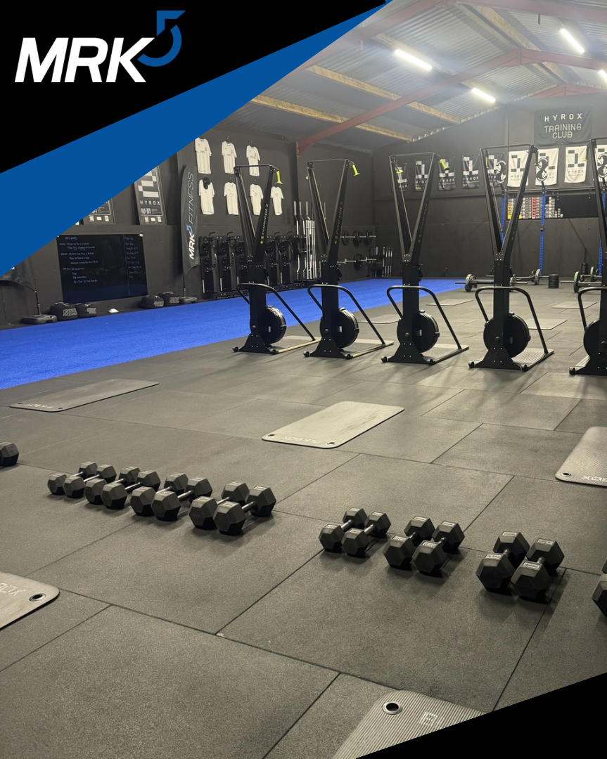 Interior of a gym with workout equipment, including dumbbells, rowing machines, squat racks, and weightlifting plates, arranged on a black rubber floor with a blue mat area and black walls.