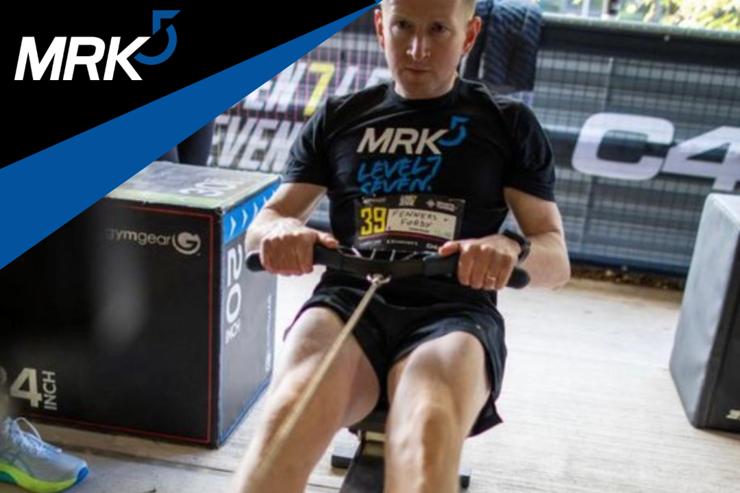 A man in a black MRK5 athletic shirt and black shorts is rowing on a rowing machine indoors. He has a focused expression, and there is a blue and black branded box and a pair of shoes nearby.