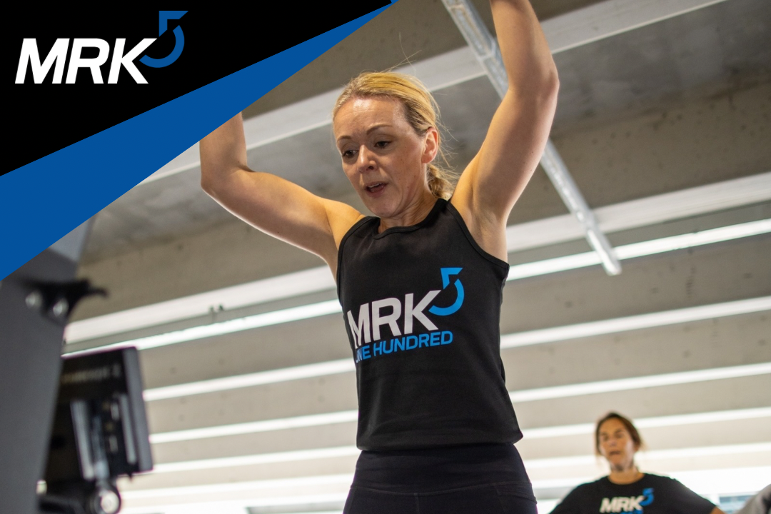 A woman lifting weights during a workout at a gym, with another woman in the background, both wearing black shirts with 'MRK5' logo.