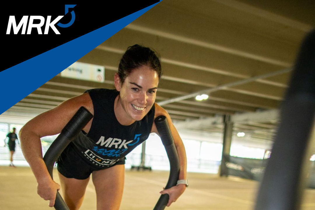 A woman smiling and pushing a sled at a fitness gym with a wooden ceiling.