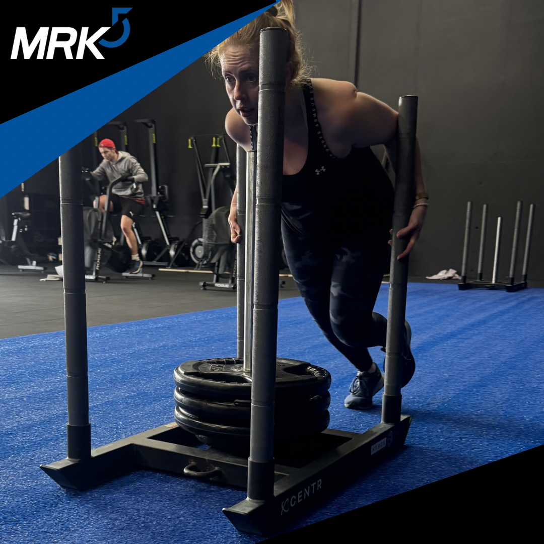A woman in workout clothes pushing a sled loaded with weights on a blue gym floor, with a man on a stationary bike in the background.