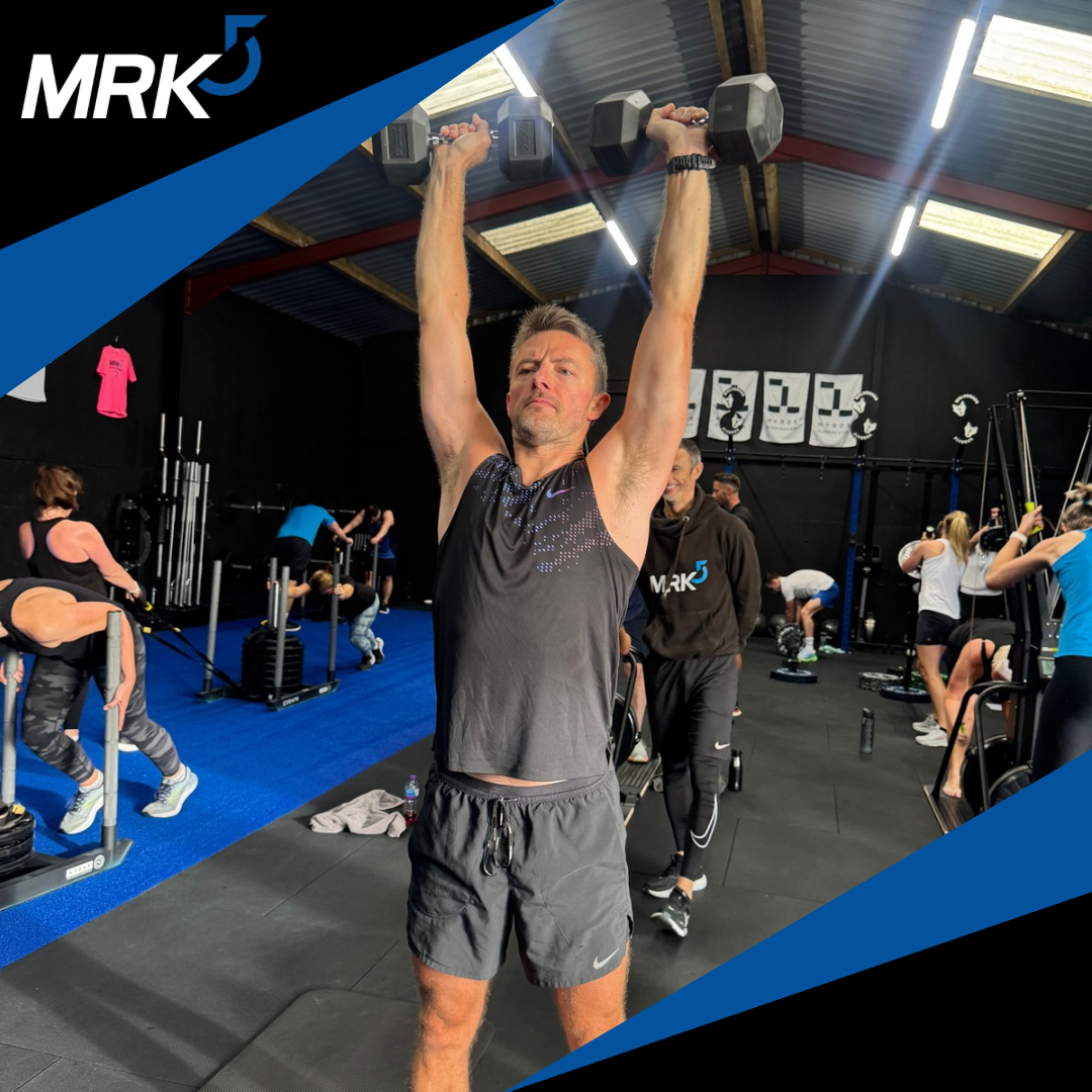 A man lifting dumbbells above his head in a gym filled with people exercising and lifting weights.