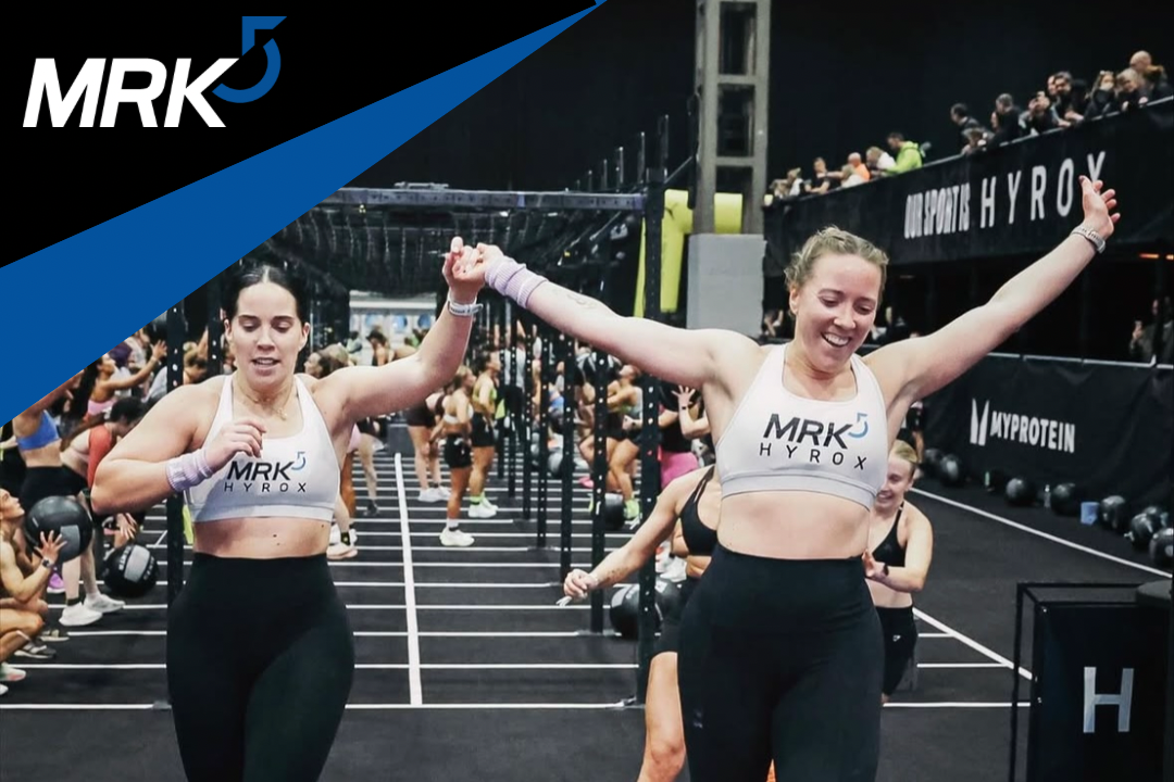 Two women in athletic wear celebrating with arms raised after a workout in a gym filled with other people exercising, fitness equipment, and black walls.