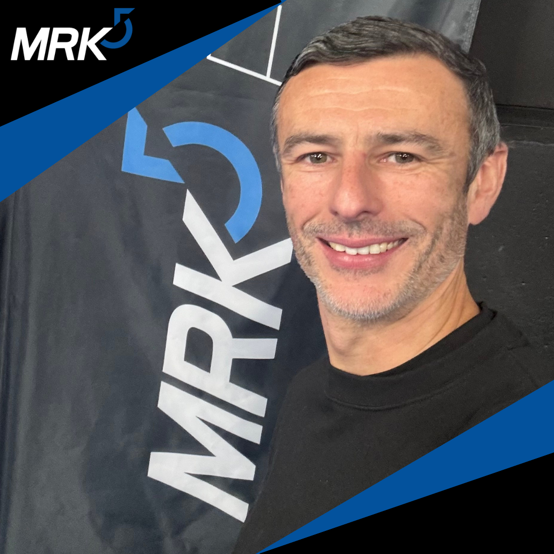 A man smiling for a selfie in front of a black wall with a logo and text reading "MRK" with blue and white accents.
