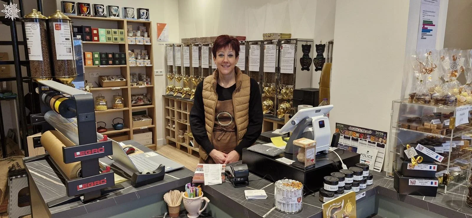 Une femme souriante derrière un comptoir dans une boutique de chocolats ou confiseries, avec des étagères remplies de produits en arrière-plan.