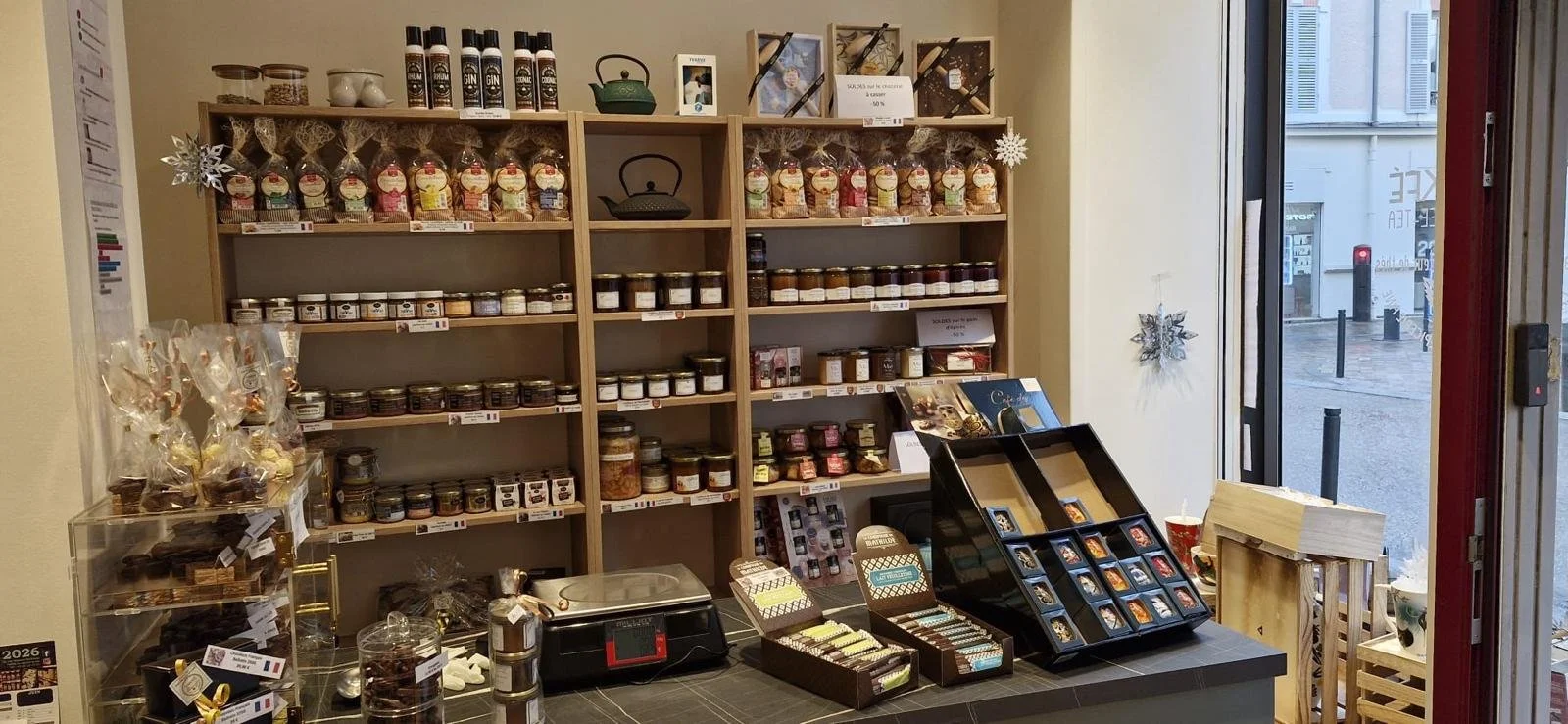 Étagère en bois avec divers produits en bocaux et emballages, face à une vitrine, avec un comptoir en dessous et une fenêtre à droite, dans un magasin de produits alimentaires.