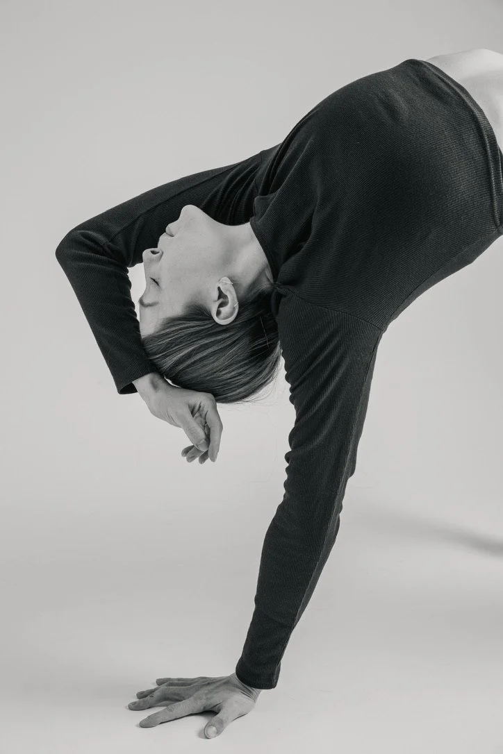 A person with short hair in a black long-sleeve shirt doing a yoga pose with one hand on the ground and the other hand behind their head, bending forward with their eyes closed.