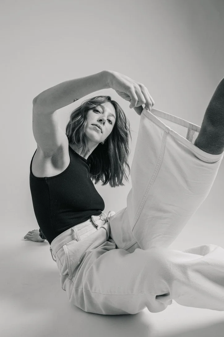 Black and white photo of woman with wavy hair, wearing a sleeveless black top and light-colored pants, sitting on the floor and holding the end of her jeans upwards.