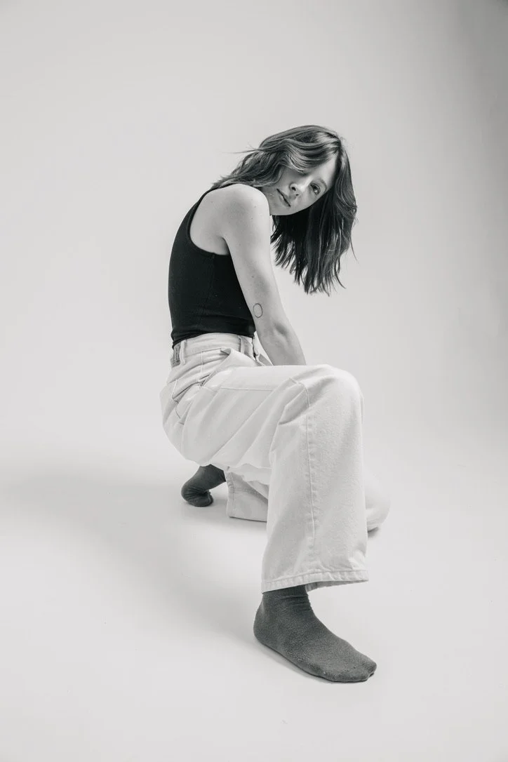 Black and white photo of a woman sitting wearing a black sleeveless top, wide-leg pants, and socks, in a minimal studio setting.