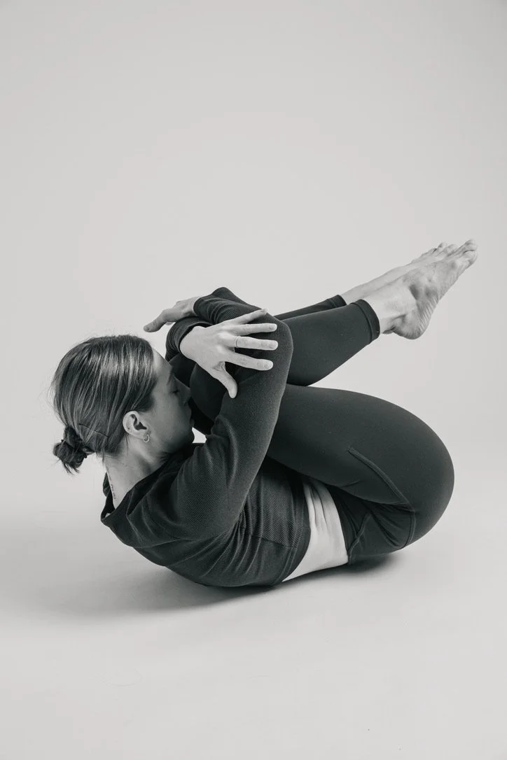 A woman performing a yoga pose on a plain background, lying on her side with her knees bent to her chest and holding her feet with her hands.