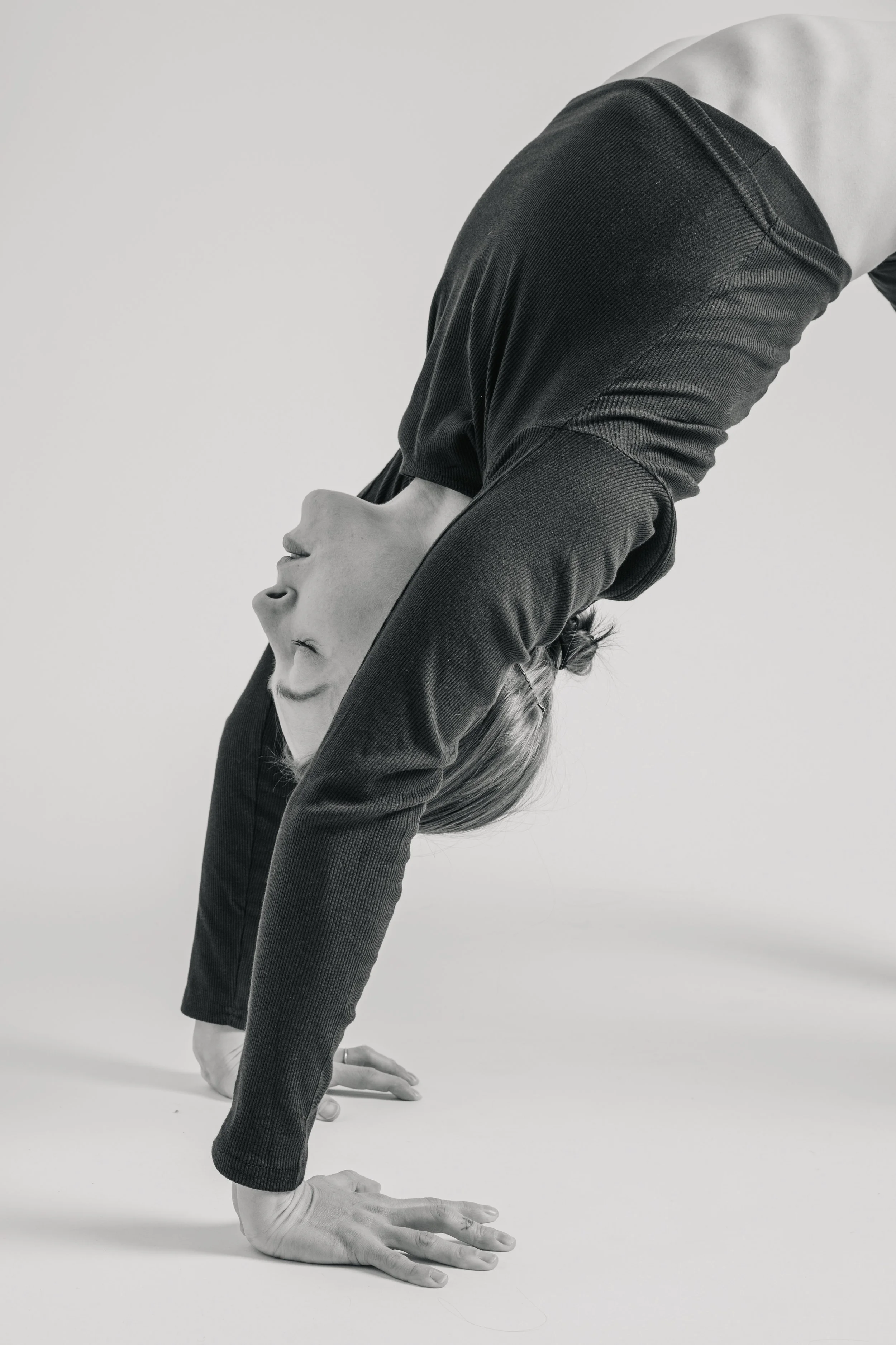 A woman in a black long-sleeve top practicing a back-bend yoga pose on a white background.