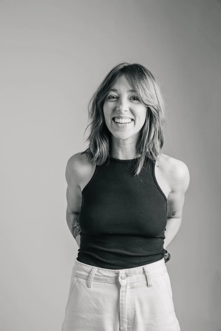Black and white photo of a woman with shoulder-length wavy hair, smiling, wearing a sleeveless black top and white pants, standing against a plain background.