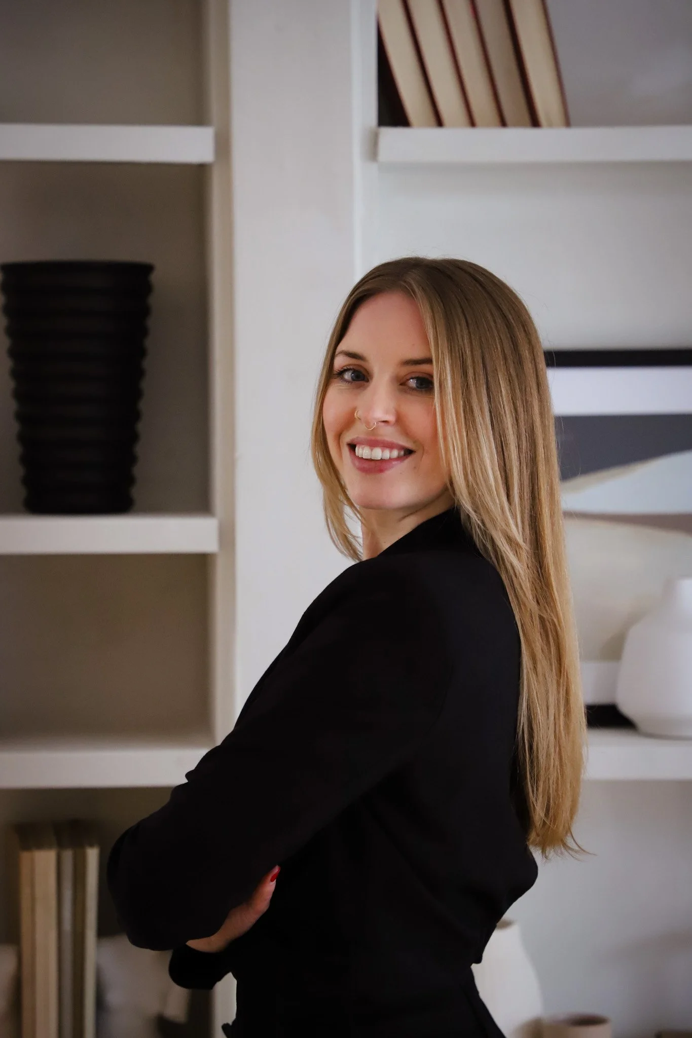 Lucy Cullen wearing a black jacket, smiling and posing with her arms crossed in front of white shelves with decorative objects.