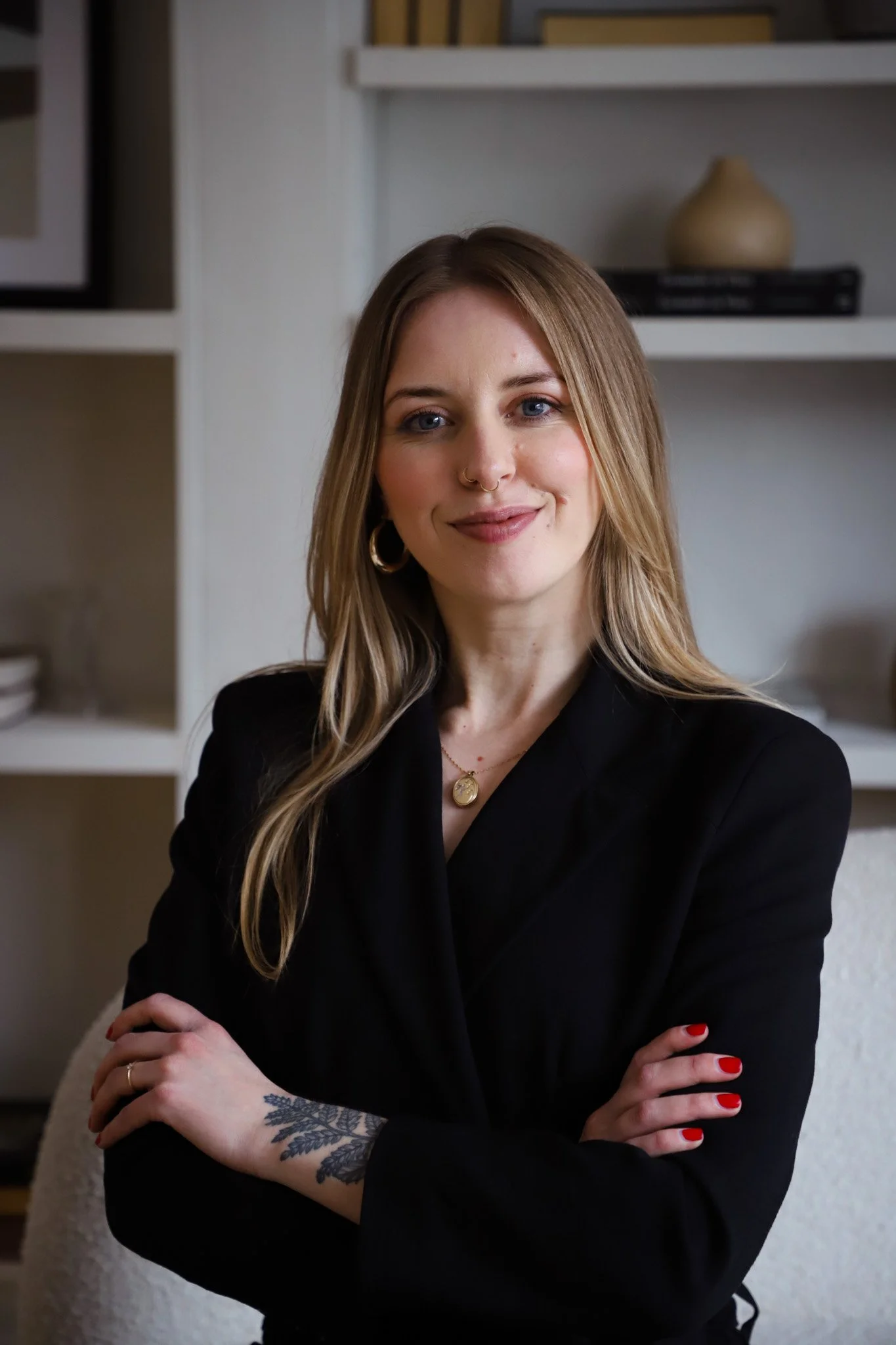 A woman with long blonde hair, blue eyes, and a nose ring, wearing a black blazer and gold jewelry, standing with crossed arms in a room with white shelves and a decorative vase.