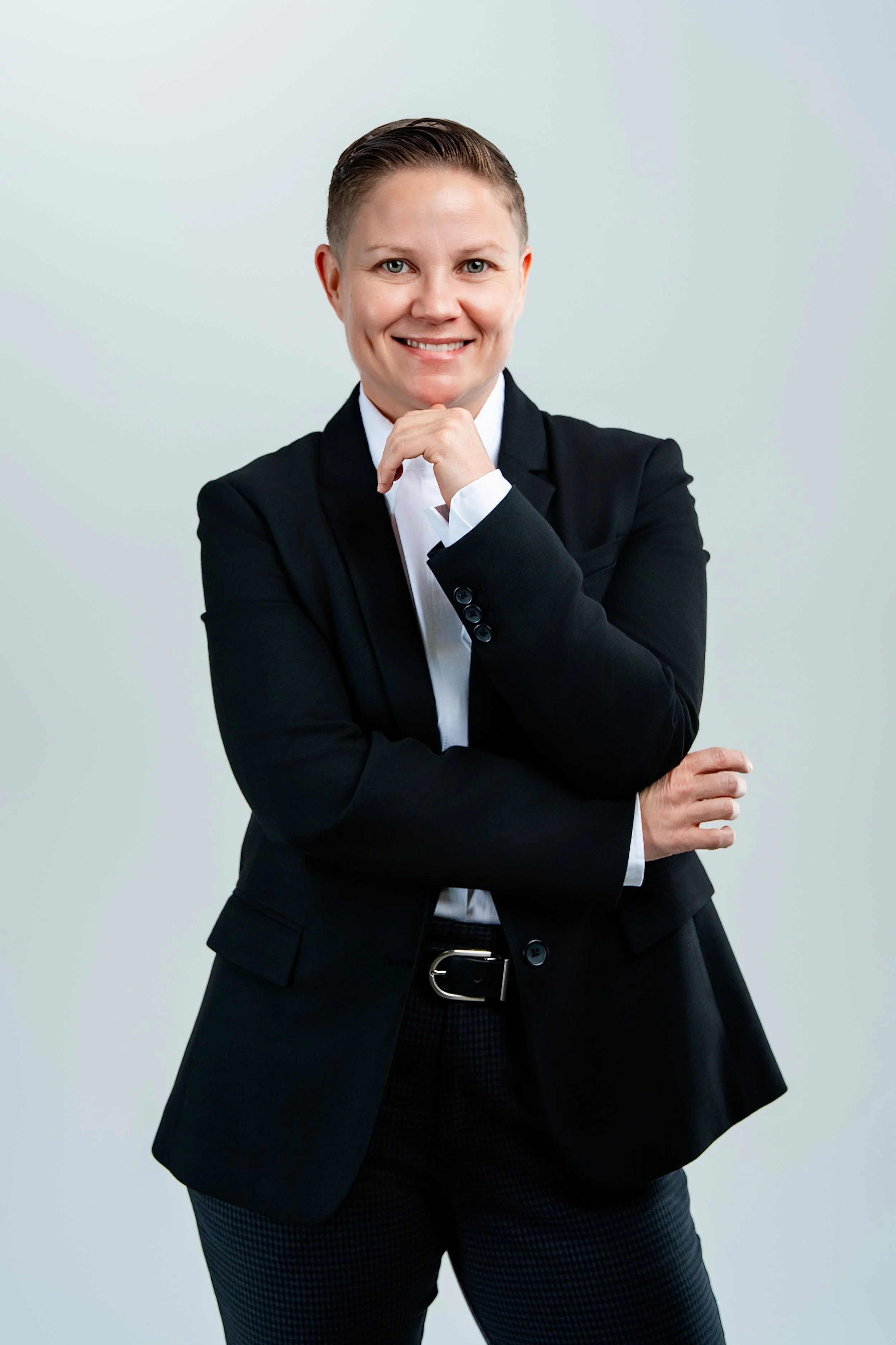 A woman with short hair wearing a black blazer, white shirt, and dark patterned pants, smiling and standing confidently with her hand resting on her chin in front of a plain light background.
