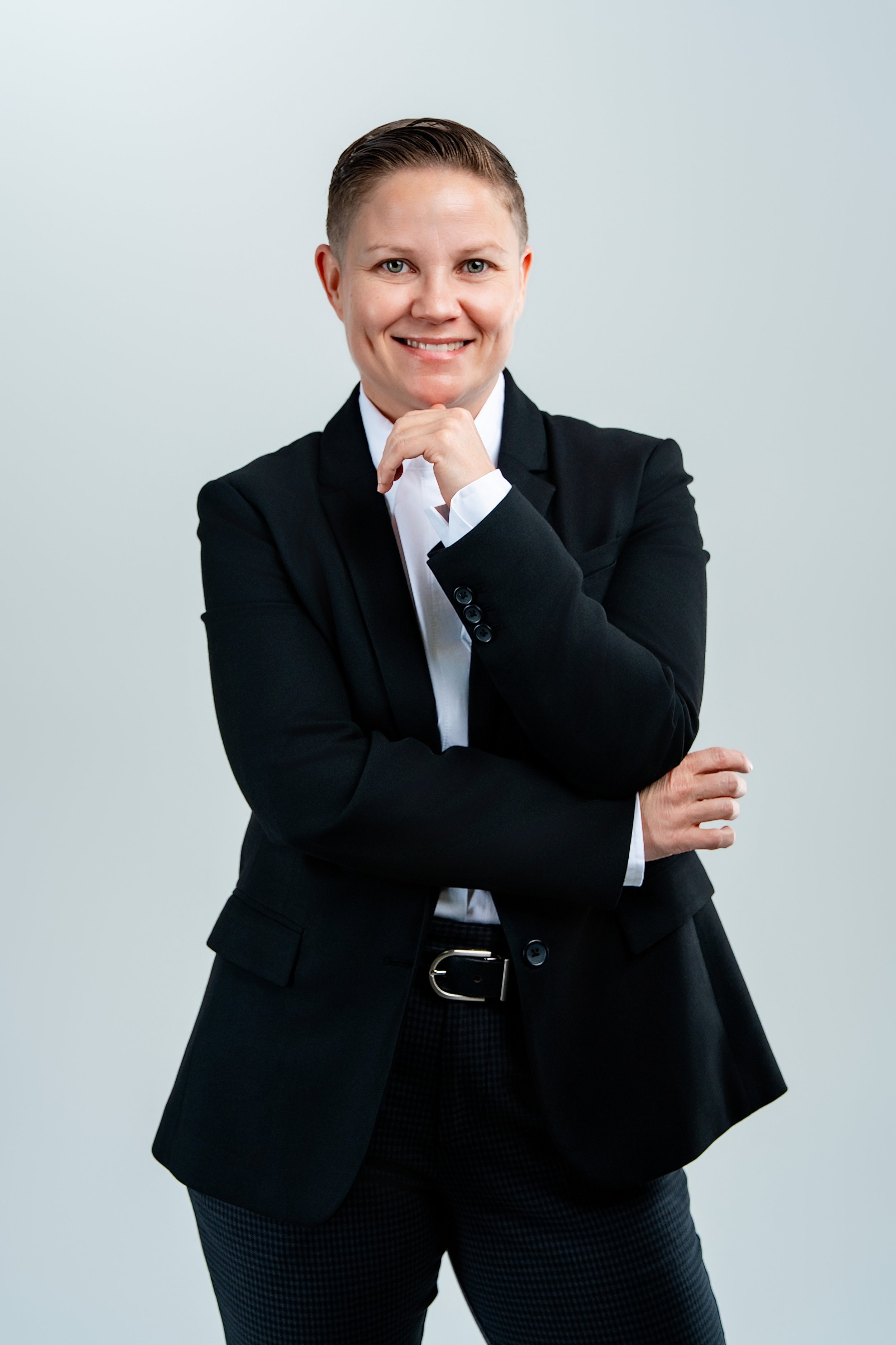 Woman with short hair wearing a black blazer and white shirt, smiling with her hand on her chin in front of a plain light gray background.