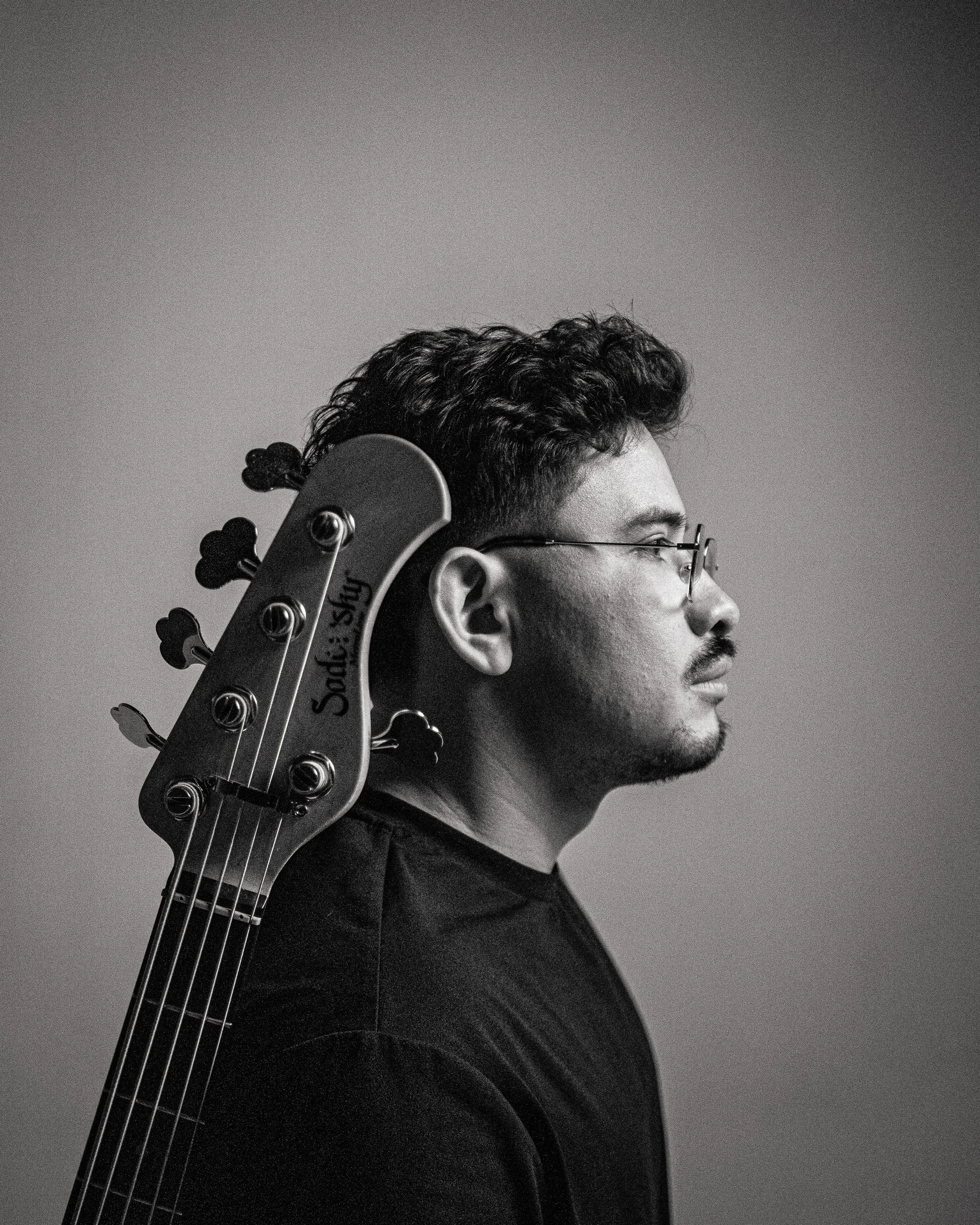 Profile of a young man with glasses, short curly hair, and a mustache, carrying a guitar on his shoulder, against a plain background.