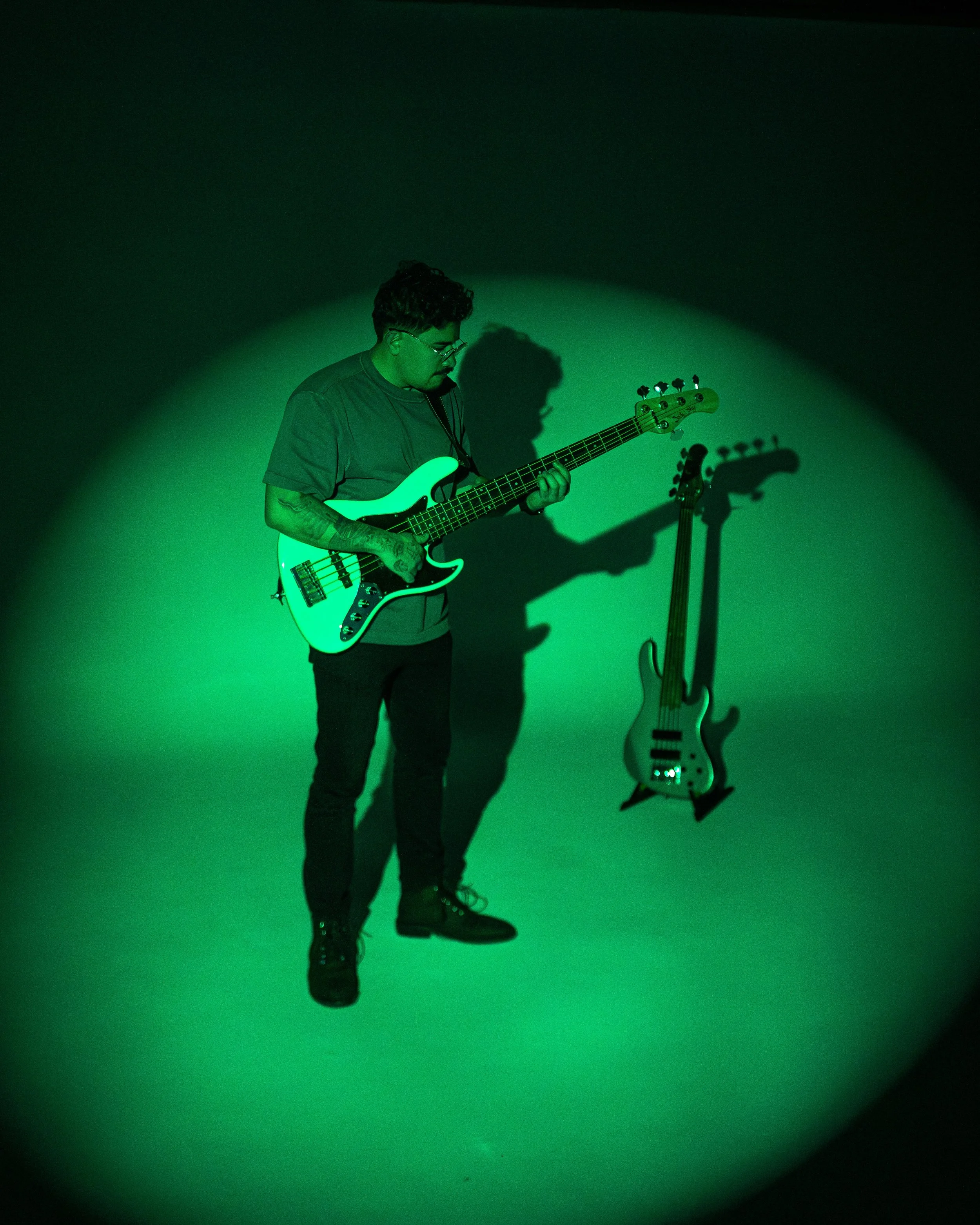 A person with curly hair, glasses, and tattoos is playing a white bass guitar. They are wearing black boots and a dark shirt, standing against a plain wall illuminated by green-tinted lighting. The person's shadow is cast on the wall.