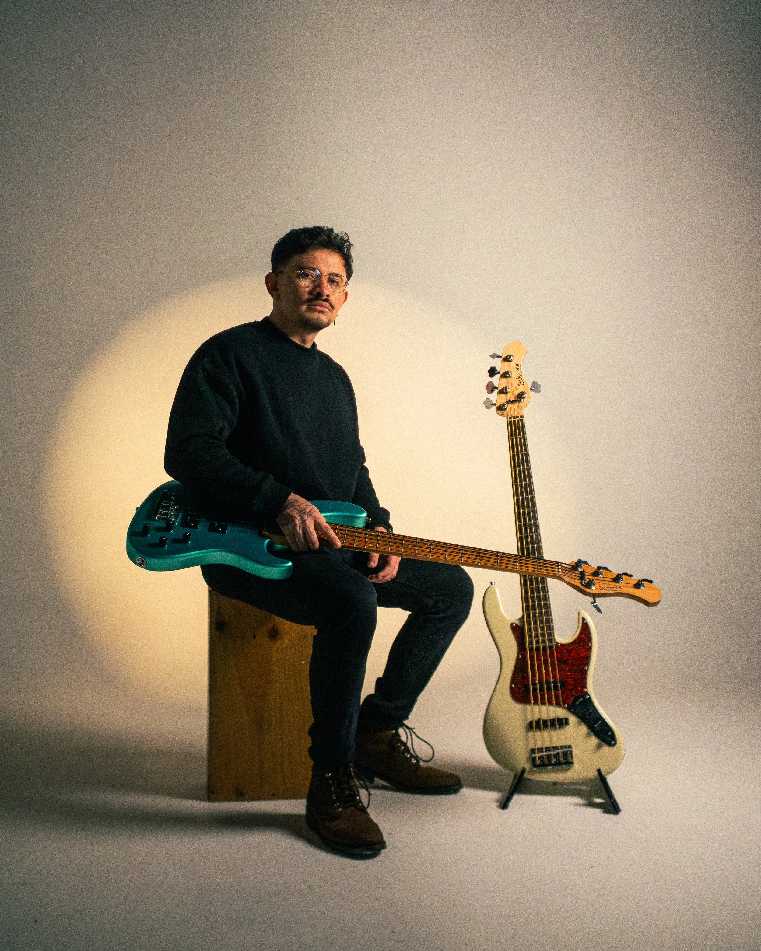 A man with glasses and a mustache sitting on a wooden box, holding a teal electric bass guitar, with two other bass guitars standing behind him, one white and one natural wood finish, against a plain off-white background.